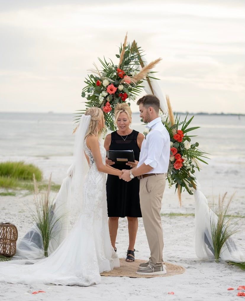 A bride and groom clasp hands under a boho pampas grass arch on the beach while officiant Beth Schell reads the ceremony.