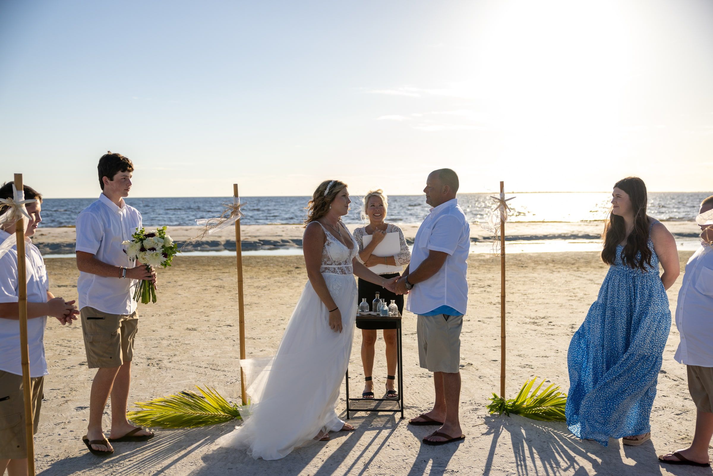A couple holds hands during their beach wedding as officiant Beth Schell smiles between them, family gathered near at sunset.