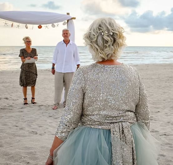 A bride in a sequined top and pale blue tulle skirt walks across the sand toward her groom, who waits under a shell-trimmed canopy with officiant Beth Schell at a sunset elopement on Sanibel.