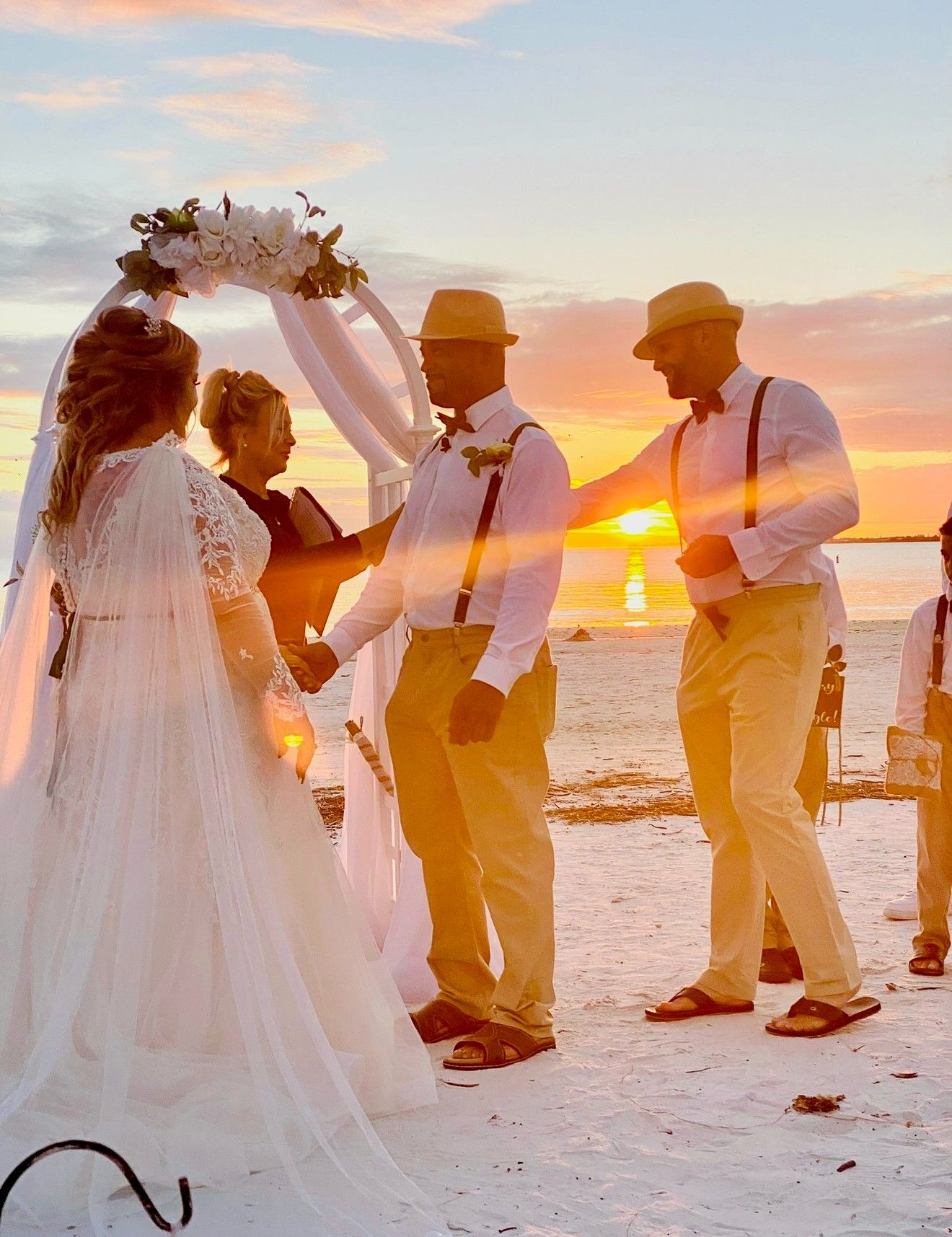A couple holds hands under a floral arch at sunset as officiant Beth Schell leads their beachside ceremony.