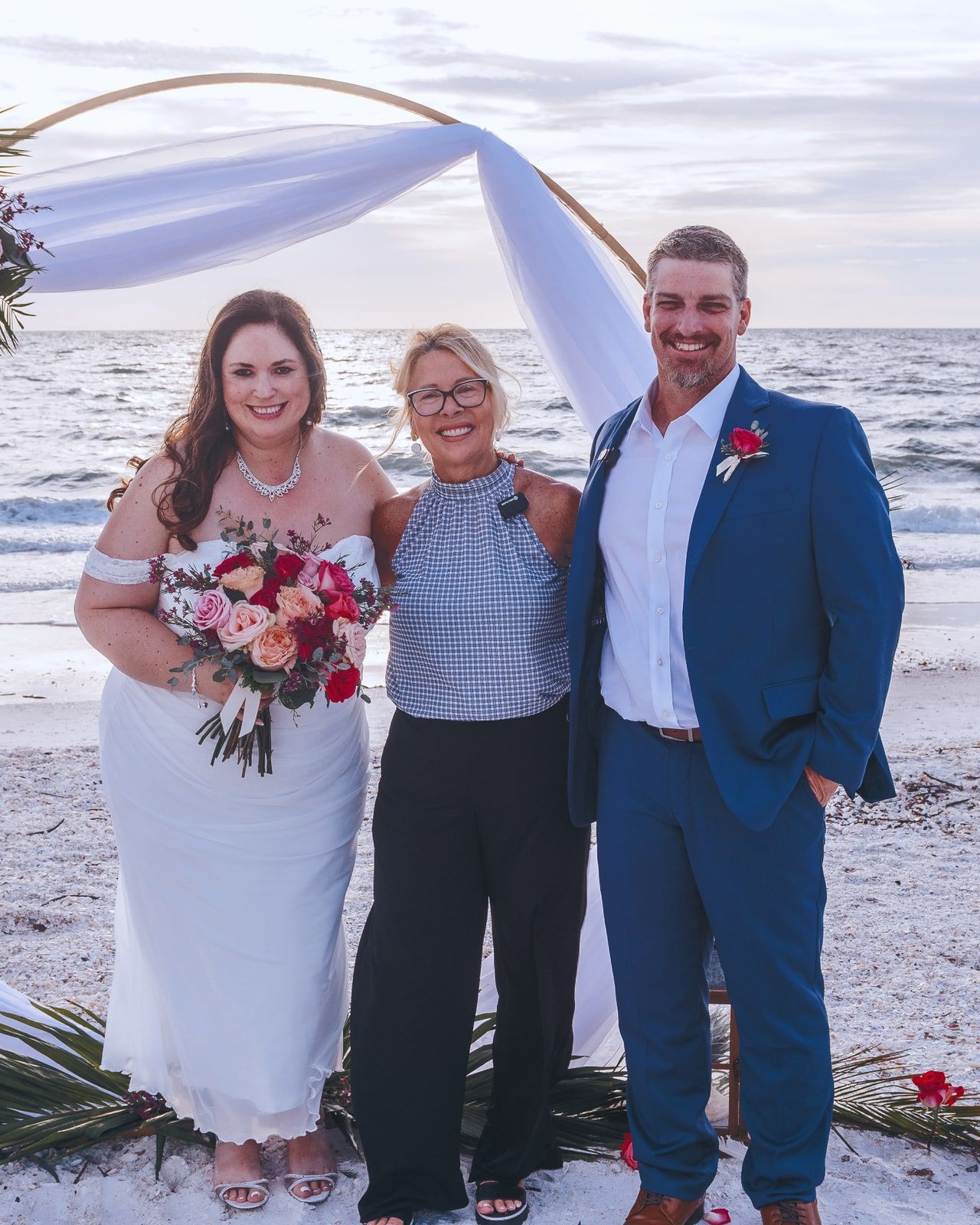 Officiant Beth Schell stands smiling with a newlywed couple beside a beachfront floral arch.